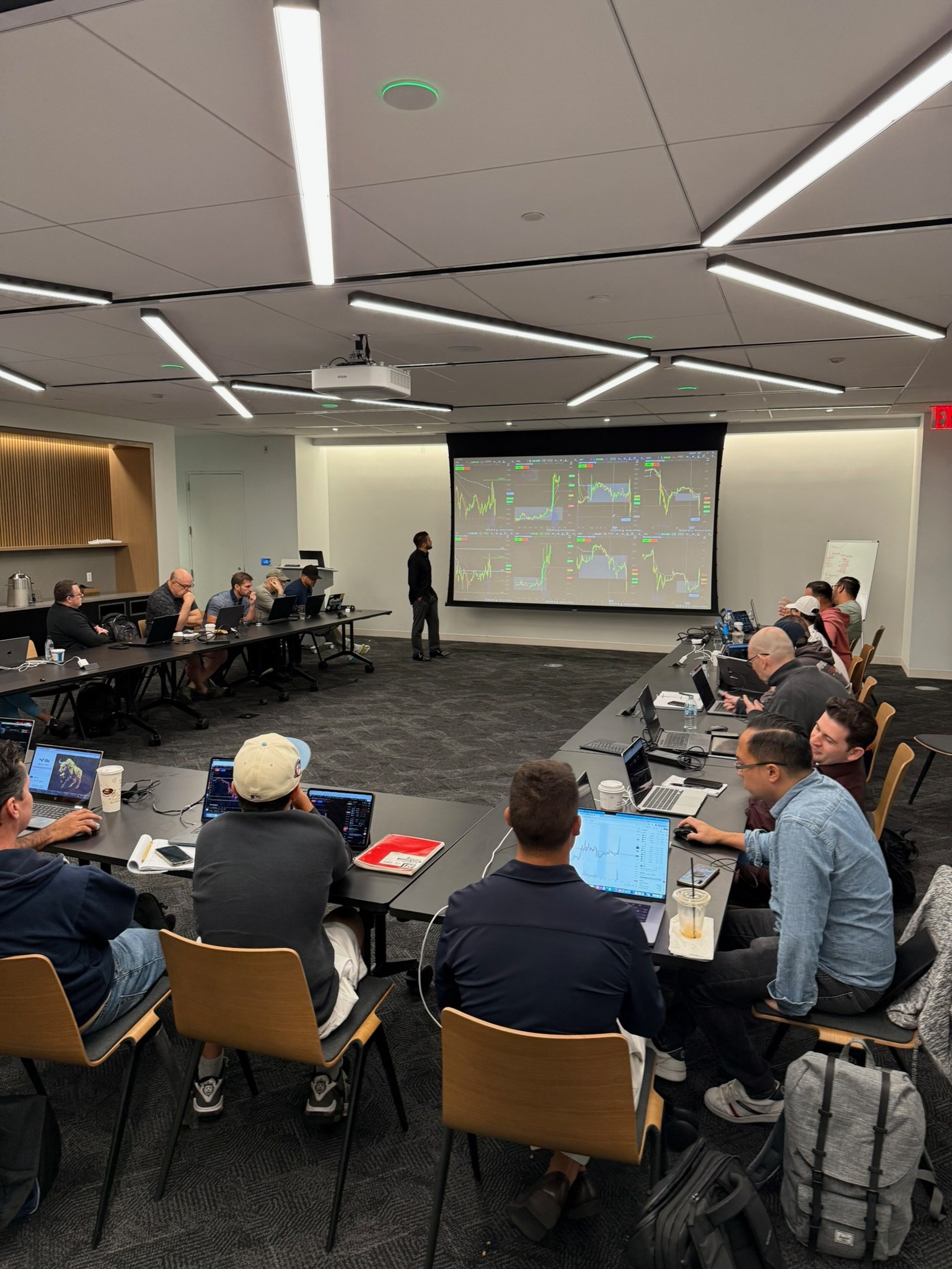 Andrew teaching a full room of students with trading charts projected on the screen while everyone works their own laptop